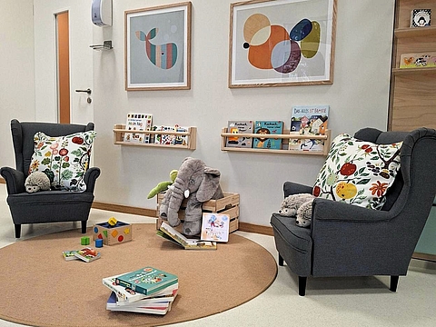 Gemütliche Leseecke in einem Fröbel-Kindergarten in Düsseldorf. Zwei graue Sessel mit bunten Kissen und Kuscheltieren stehen auf einem runden Teppich. Dazwischen liegen und stehen Kinderbücher und Holzspielzeug. An der Wand hängen zwei abstrakte Bilder sowie kleine Bücherregale mit weiteren Kinderbüchern. Die Atmosphäre wirkt freundlich, hell und einladend.